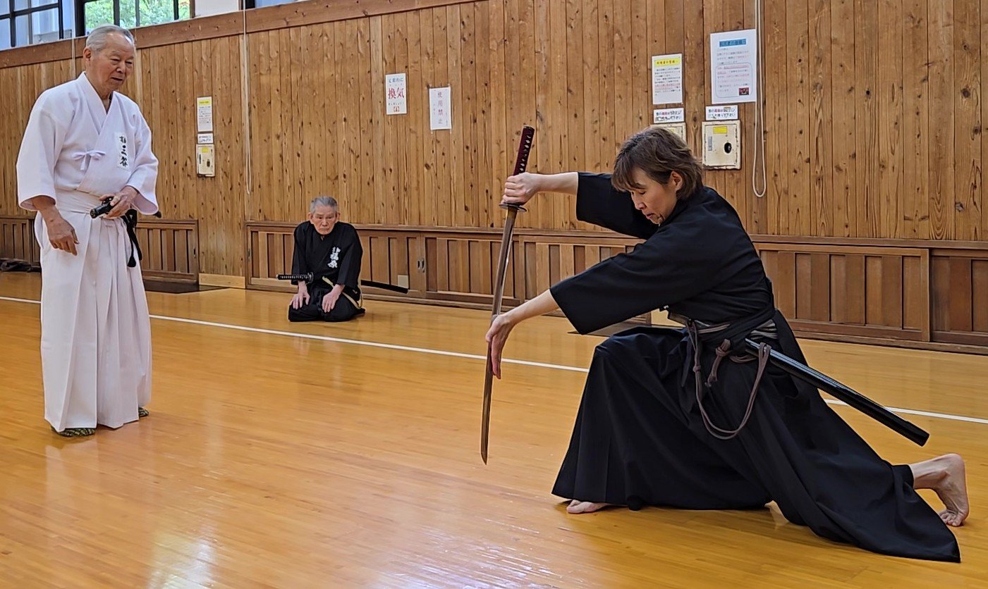 Kneeling iaidō practitioner holds a drawn sword vertically while two other practitioners observe.