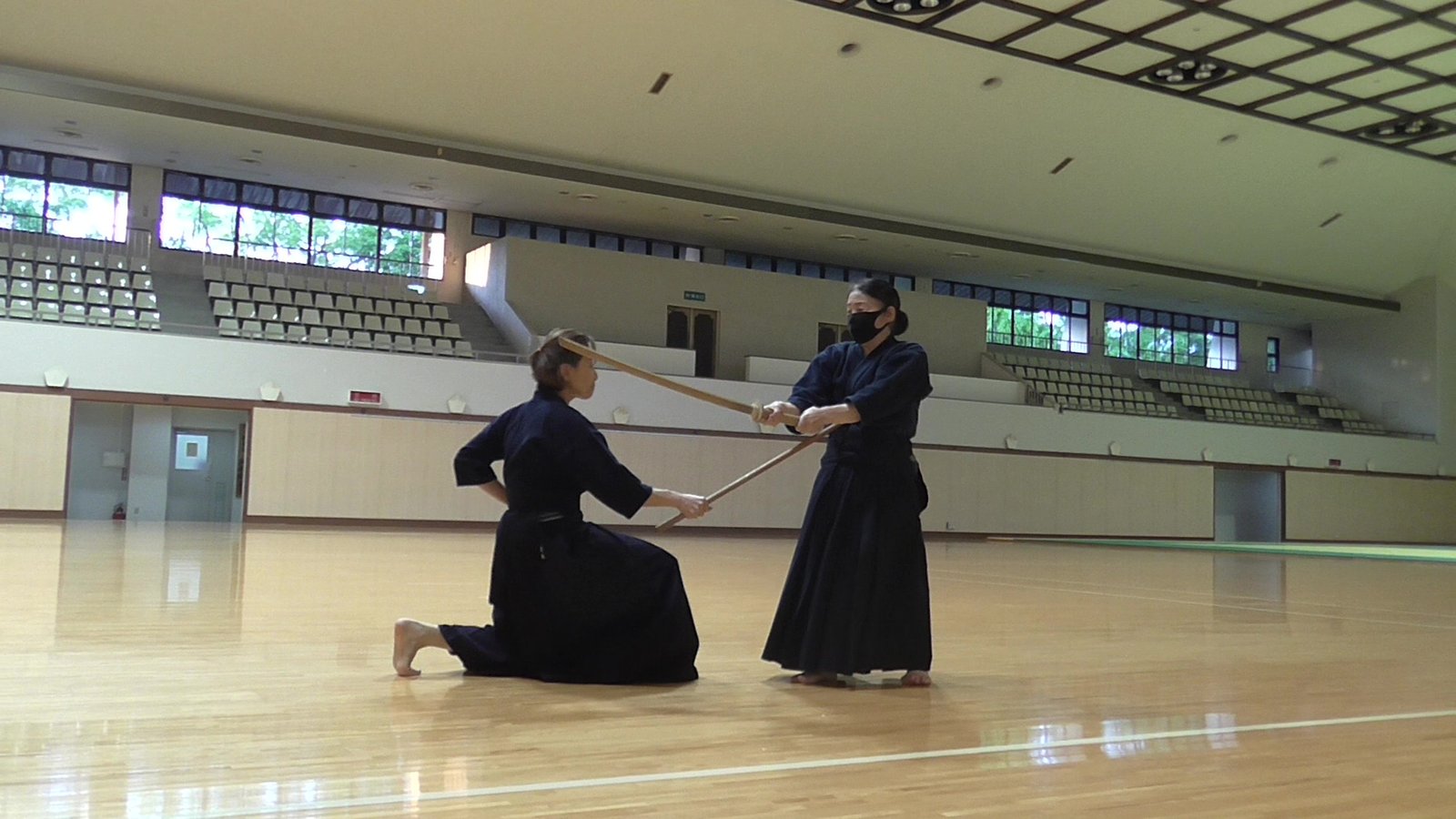 Kneeling jōdō practitioner uses a wooden staff against a standing partner holding a wooden sword.