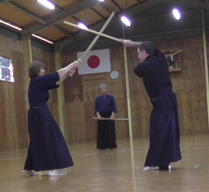 Two jōdō practitioners cross a wooden staff and sword overhead as another practitioner watches.
