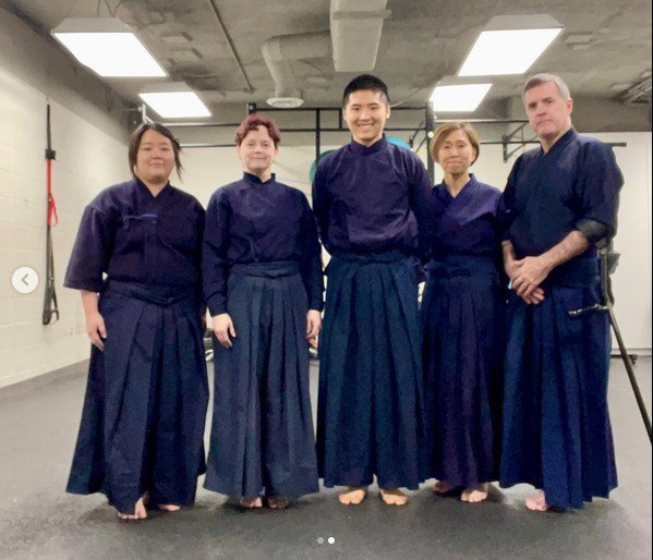 Five practitioners in dark uniforms and hakama stand together for a group portrait.